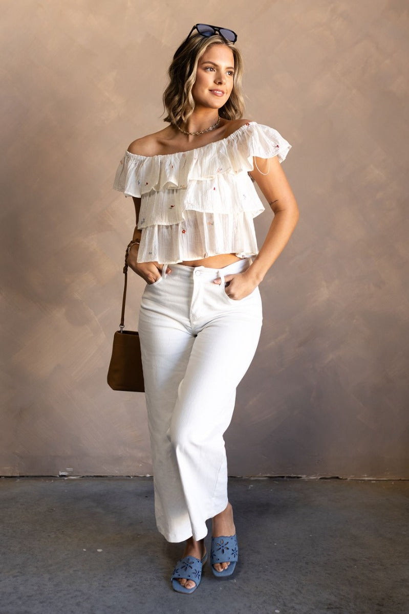 Full body front view of model wearing the Carolina Ivory Tiered Floral Top that has ivory light weight fabric, a three tiered design, multi color embroidered flower details, a square neckline and ruffle straps.