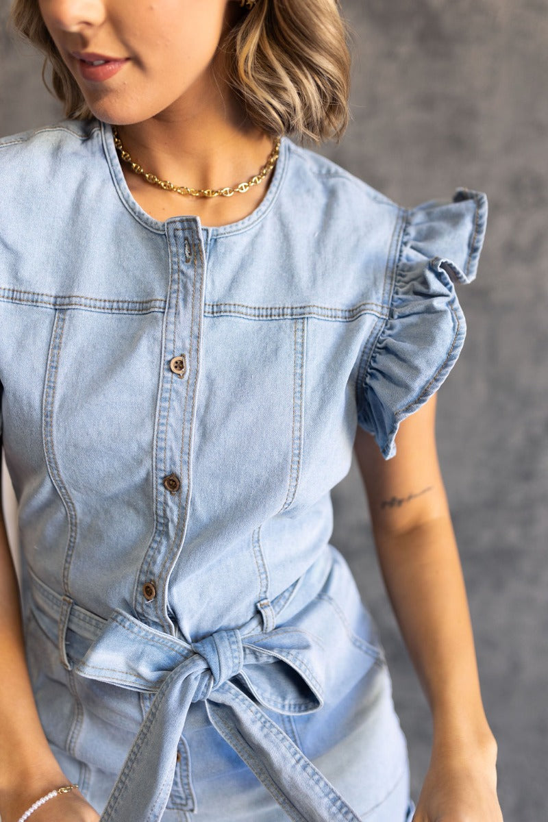 CLose up view of model wearing the Ruth Light Denim Ruffle Short Sleeve Romper which features light wash denim fabric, light brown stitch details, folded hem, two front pockets, belt loops with tie belt, wooden button up front closure, round neckline and 