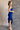 Side view of model wearing the Liliana Blue Asymmetrical Sleeveless Midi Dress which features royal blue knit fabric, an asymetrical ruffle hem, midi length, a square neckline, and adjustable straps.