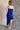 Full body back view of model wearing the Liliana Blue Asymmetrical Sleeveless Midi Dress which features royal blue knit fabric, an asymetrical ruffle hem, midi length, a square neckline, and adjustable straps.