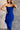 Close up view of model wearing the Liliana Blue Asymmetrical Sleeveless Midi Dress which features royal blue knit fabric, an asymetrical ruffle hem, midi length, a square neckline, and adjustable straps.