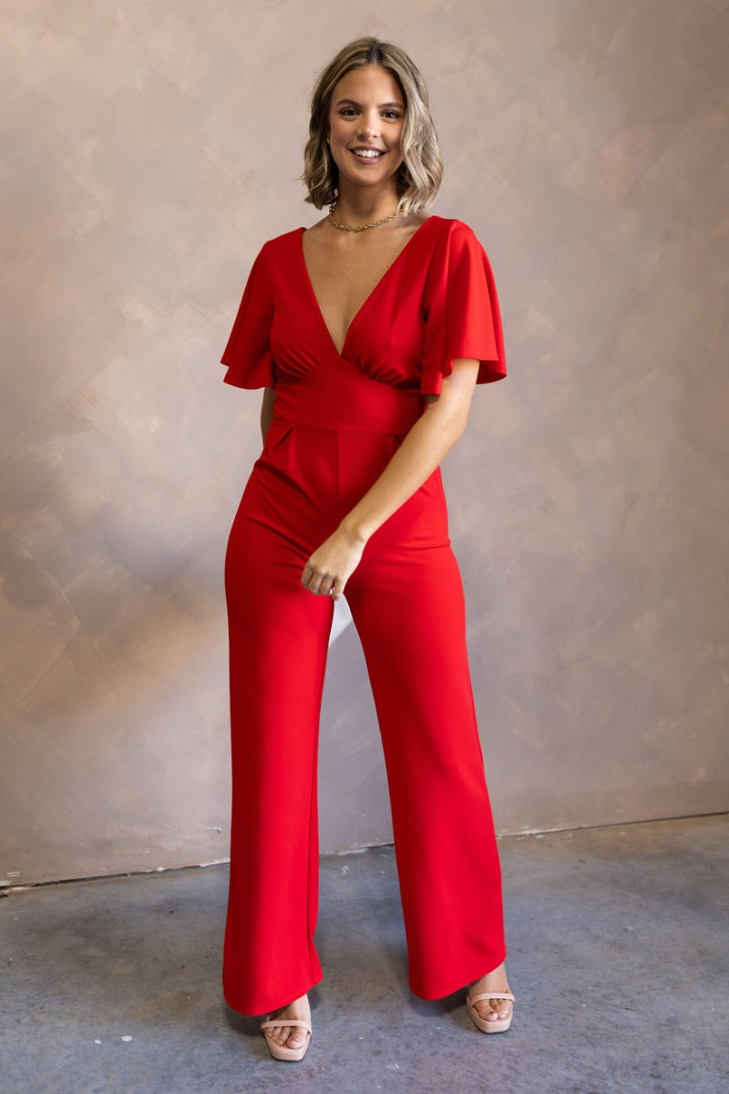 Full body view of model wearing the Jessica Red Plunge Neckline Jumpsuit which features red knit fabric, a plunge neckline, short flare sleeves, an open back, a monochrome back zipper with a hook closure, and flare pant legs.