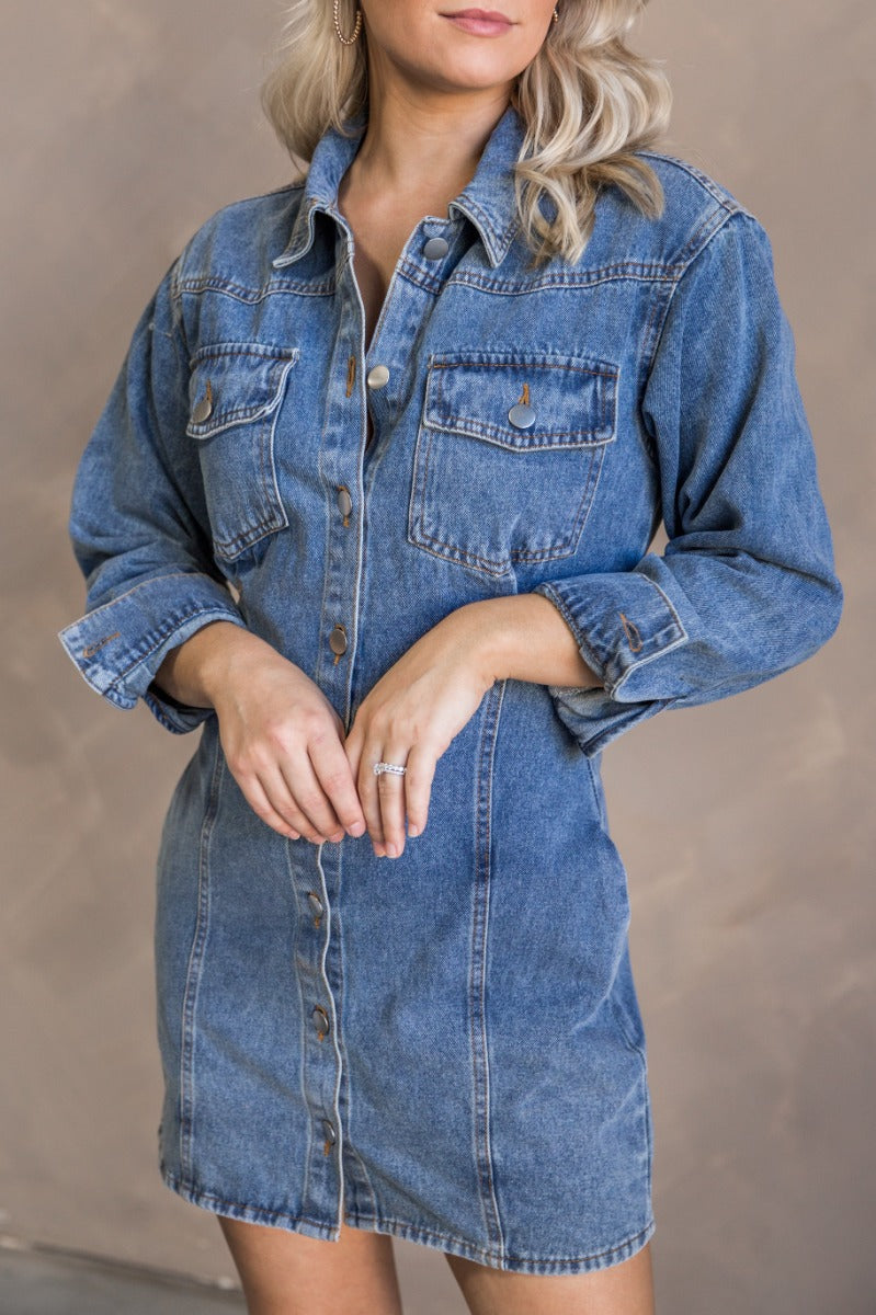 Close up view of model wearing the Tessa Blue Denim Long Sleeve Mini Dress which features blue denim fabric, front button up, collared neckline, two front chest buttoned pockets, brown stitching details and long sleeves with buttoned cuffs.