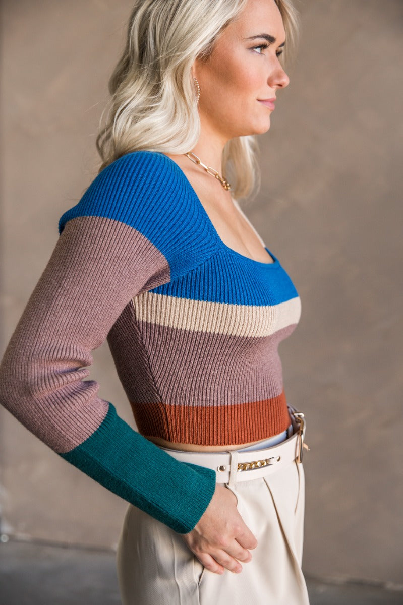 Side view of model wearing the Natalie Blue Multi Cropped Long Sleeve Top which features taupe, rust, beige and blue knit fabric, striped pattern, cropped waist, square neckline and long sleeves.