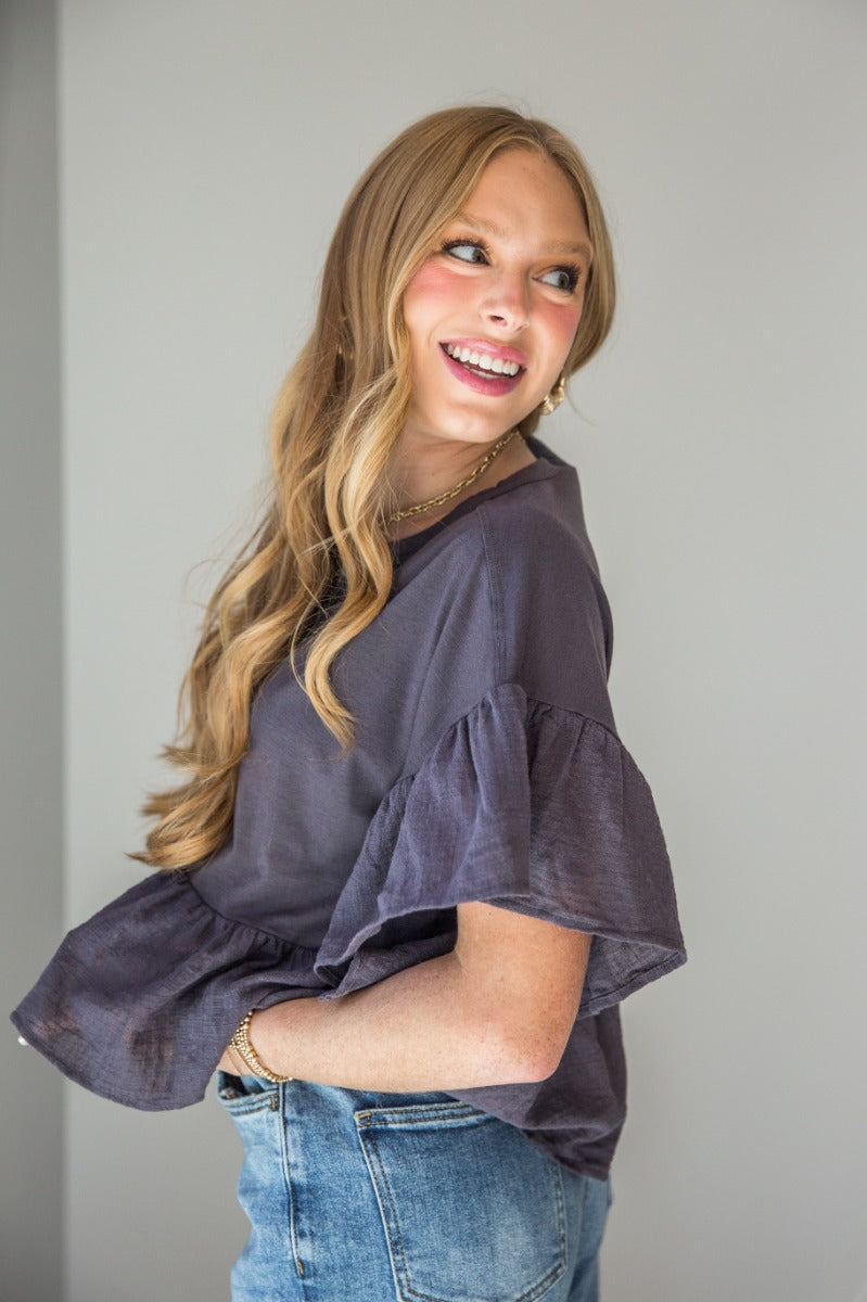 Side view of model wearing the Alyssa Charcoal Ruffled Short Sleeve Top that has charcoal knit fabric, ruffled guaze short sleeves, a gauze peplum bottom, and a notched neckline.