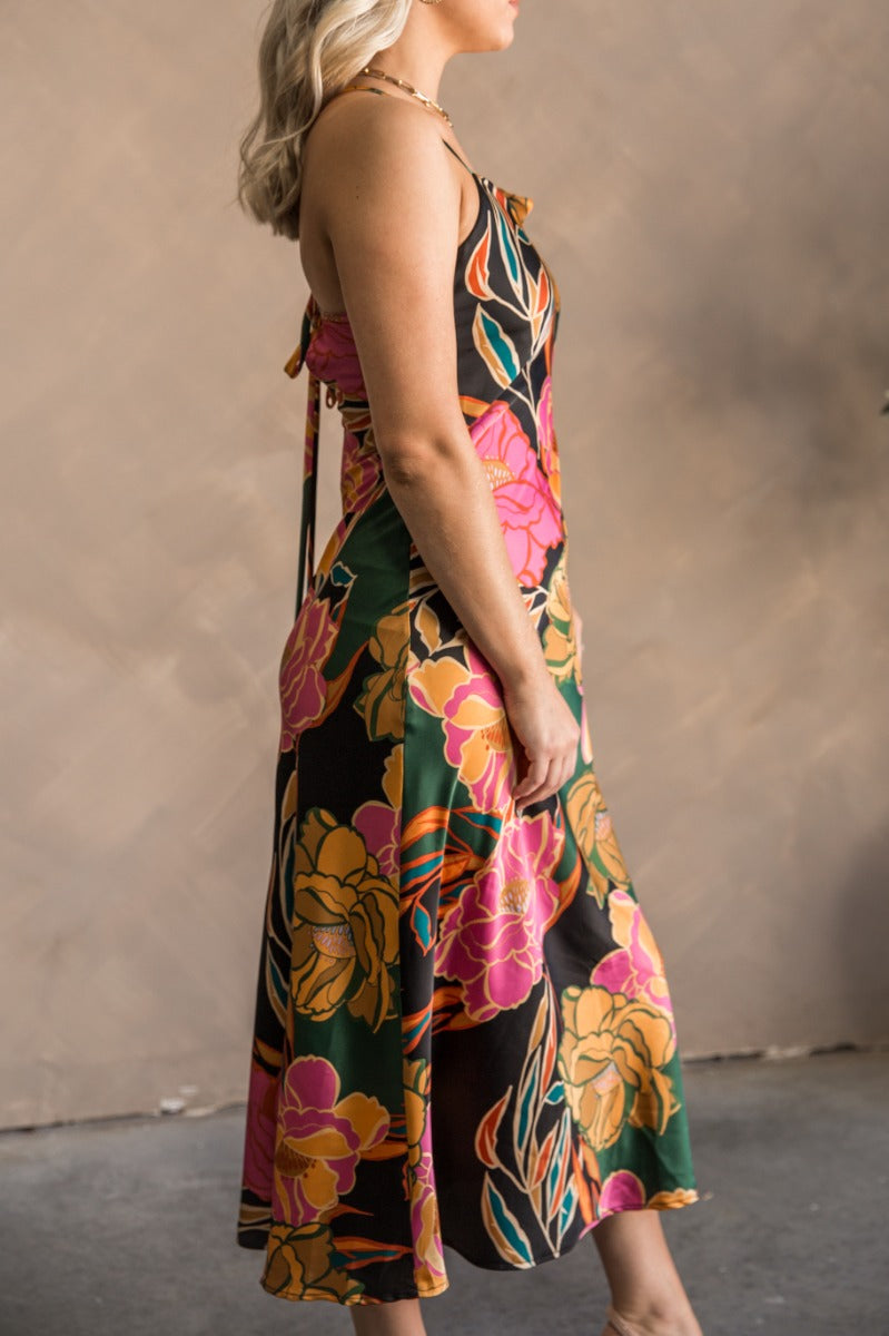 Side view of model wearing the Elise Multi Floral Satin Midi Dress which features black, green, pink, orange, turquoise, beige and dark yellow satin fabric, floral print, midi length, cowl neckline, adjustable straps, corset back ties and sleeveless.