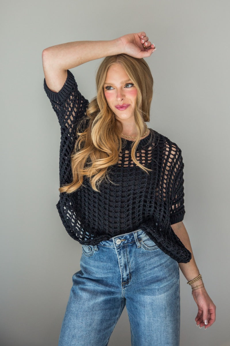 Front view of model wearing the Tatum Black Open Knit Short Sleeve Top which features black crochet knit fabric, round neckline and short sleeves.