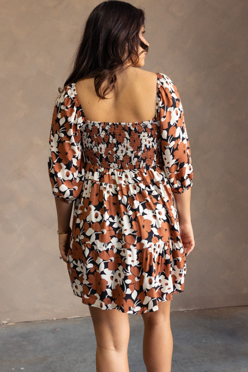 back view of model wearing the Selah Black & Brown Floral Mini Dress that has a black, brown and white floral print, a flare skirt,a scooped neckline, short puff sleeves, and a smocked back.