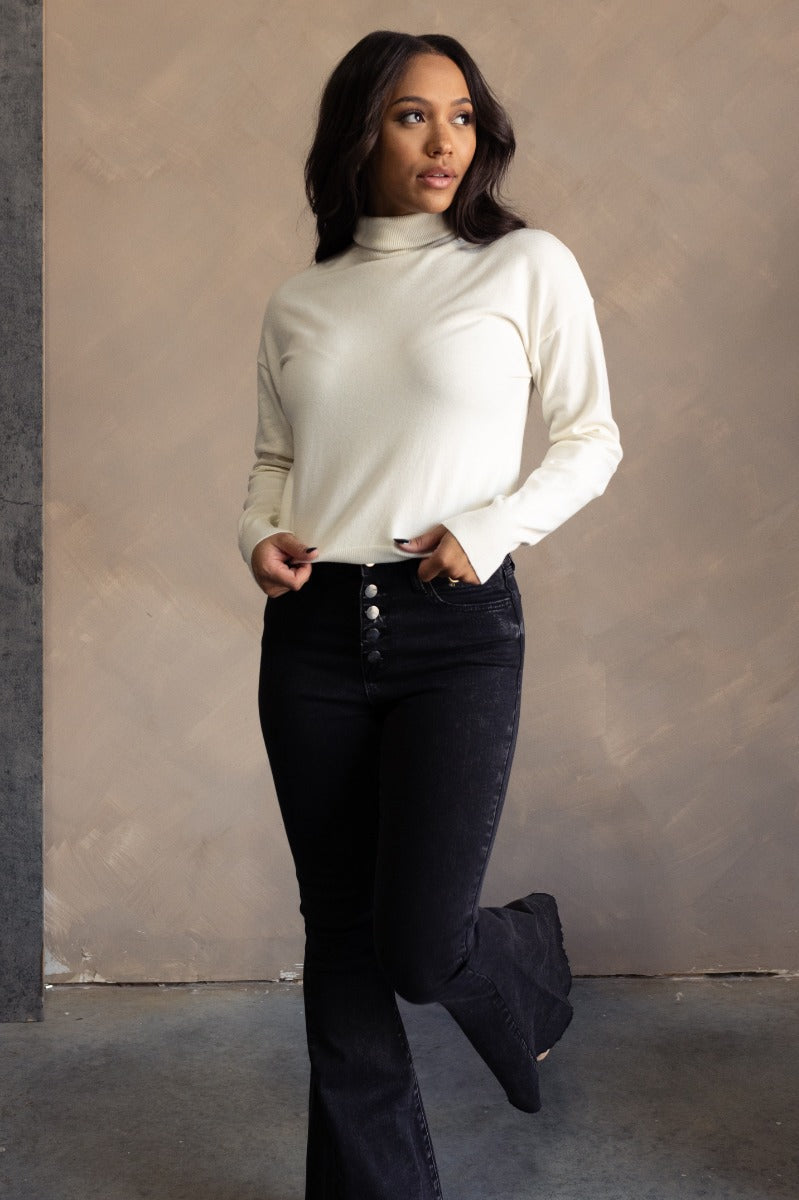Full body view of model wearing the Gabriela Cream Turtleneck Sweater which features cream knit fabric, ribbed trim, a turtleneck neckline, dropped shoulders, and long sleeves with cuffs.
