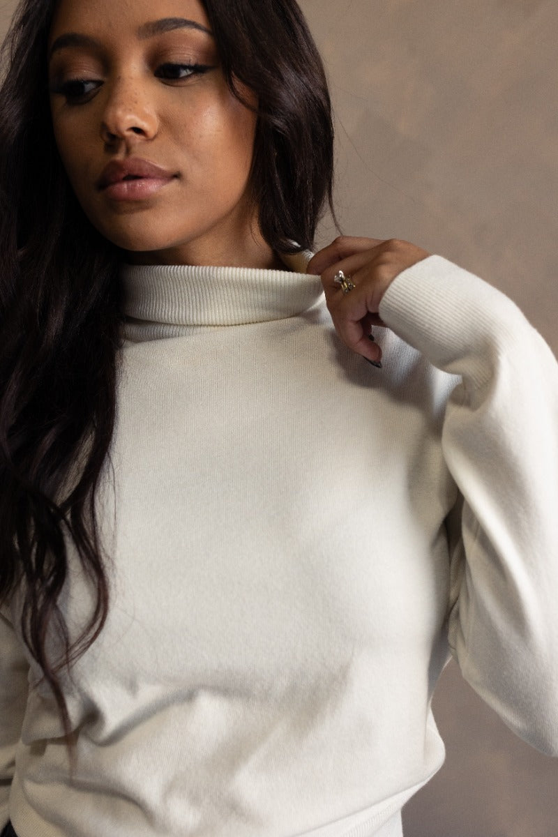 Close up view of model wearing the Gabriela Cream Turtleneck Sweater which features cream knit fabric, ribbed trim, a turtleneck neckline, dropped shoulders, and long sleeves with cuffs.