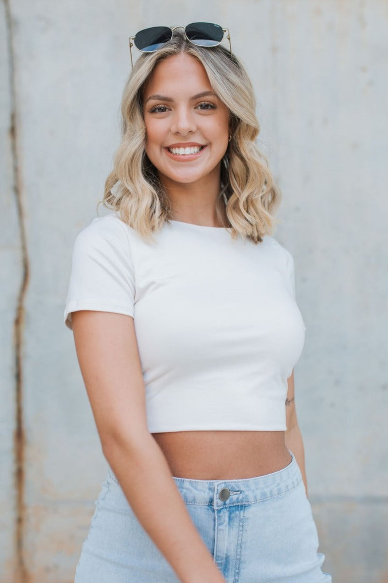 Close front view of model wearing the Play It Cool Crop Top that features ivory fabric, a cropped waist, a round neckline and short sleeves.