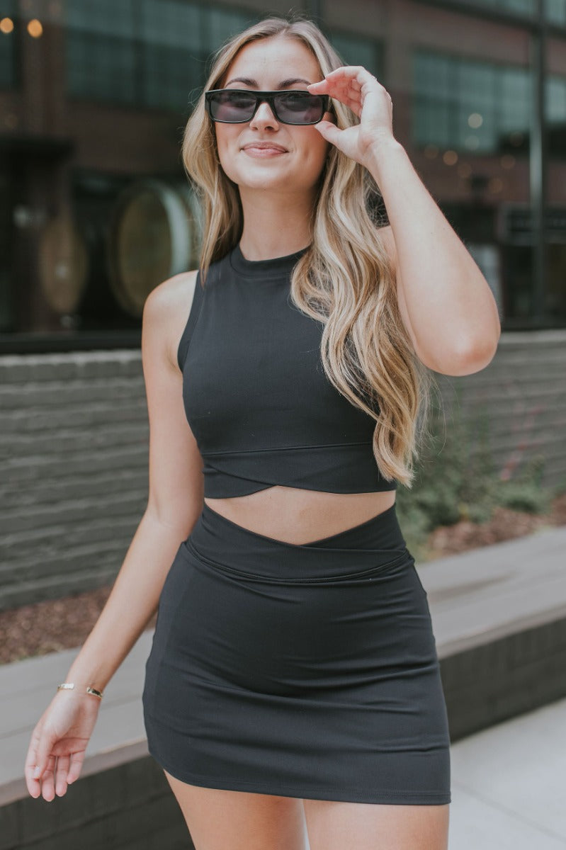 Front view of model wearing the Get Obsessed Tank in Black that has black fabric, a cropped waist, a high round neckline, bra padding, and a sleeveless design