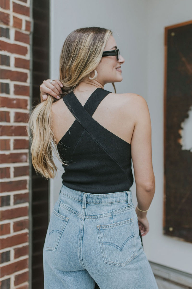 Back  view of model wearing the Weekend Lover Tank which features black fabric, a cropped waist, a halter neckline, and criss cross straps.