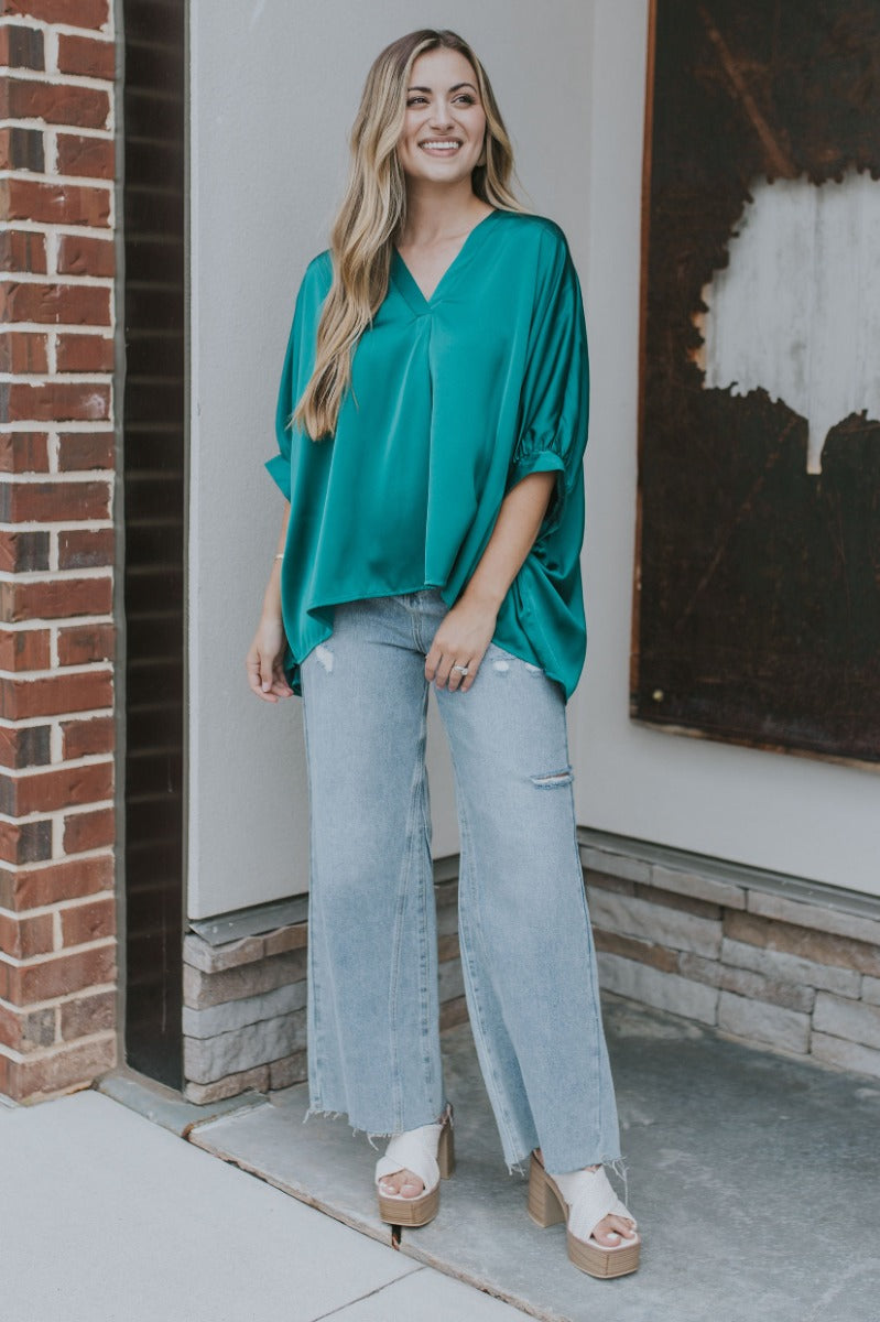 Full body view of model wearing the Some Kind Of Love Top in Green which features emerald green satin fabric, a high low hem, a v-neckline and half puff sleeves.