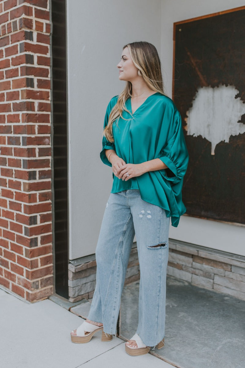 Side view of model wearing the Some Kind Of Love Top in Green which features emerald green satin fabric, a high low hem, a v-neckline and half puff sleeves.