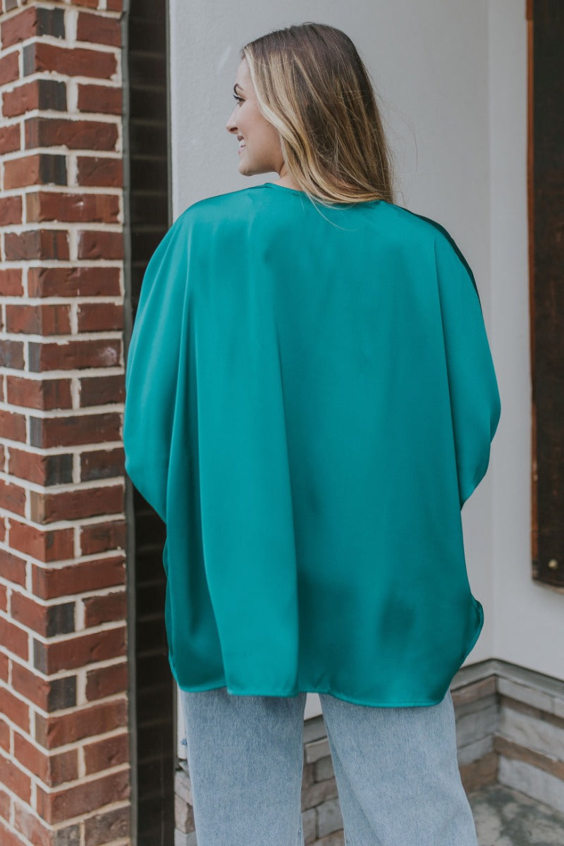 Back view of model wearing the Some Kind Of Love Top in Green which features emerald green satin fabric, a high low hem, a v-neckline and half puff sleeves.