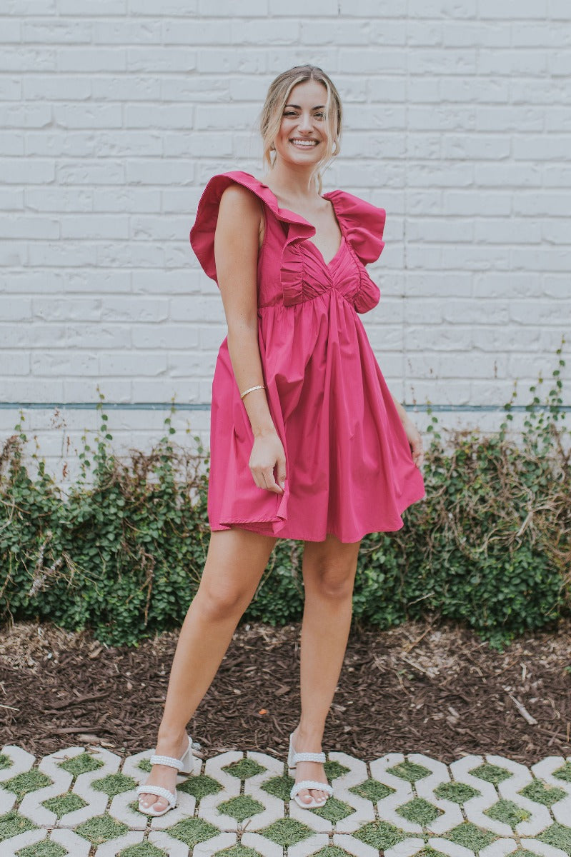 Full body view of model wearing the Total Knockout Dress in Pink which features magenta fabric, mini length, a v-neckline, side pockets, pleated upper details, dramatic ruffle straps, magenta lining, a smocked upper, and a monochromatic back zipper with a