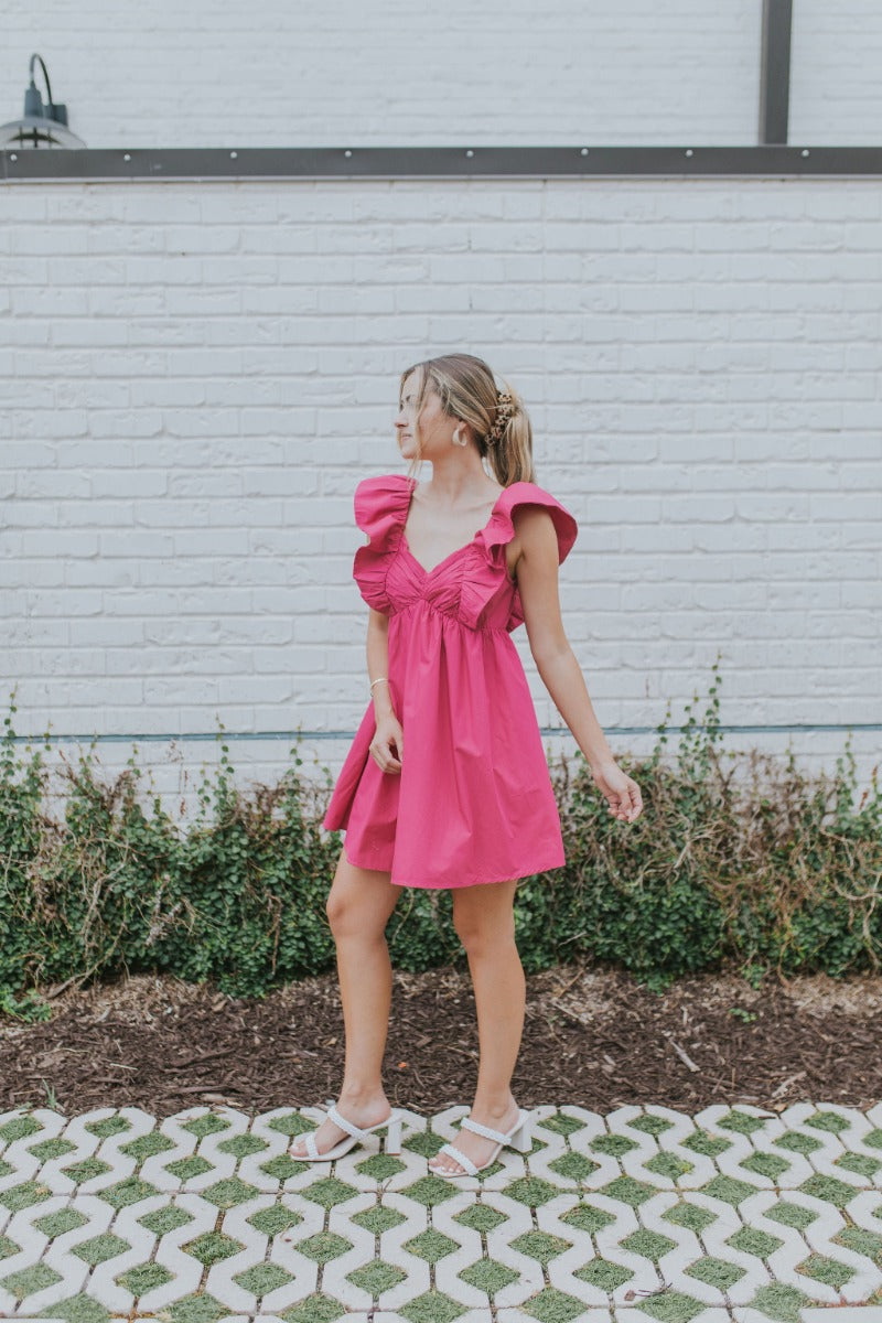 Full body side  view of model wearing the Total Knockout Dress in Pink which features magenta fabric, mini length, a v-neckline, side pockets, pleated upper details, dramatic ruffle straps, magenta lining, a smocked upper, and a monochromatic back zipper 