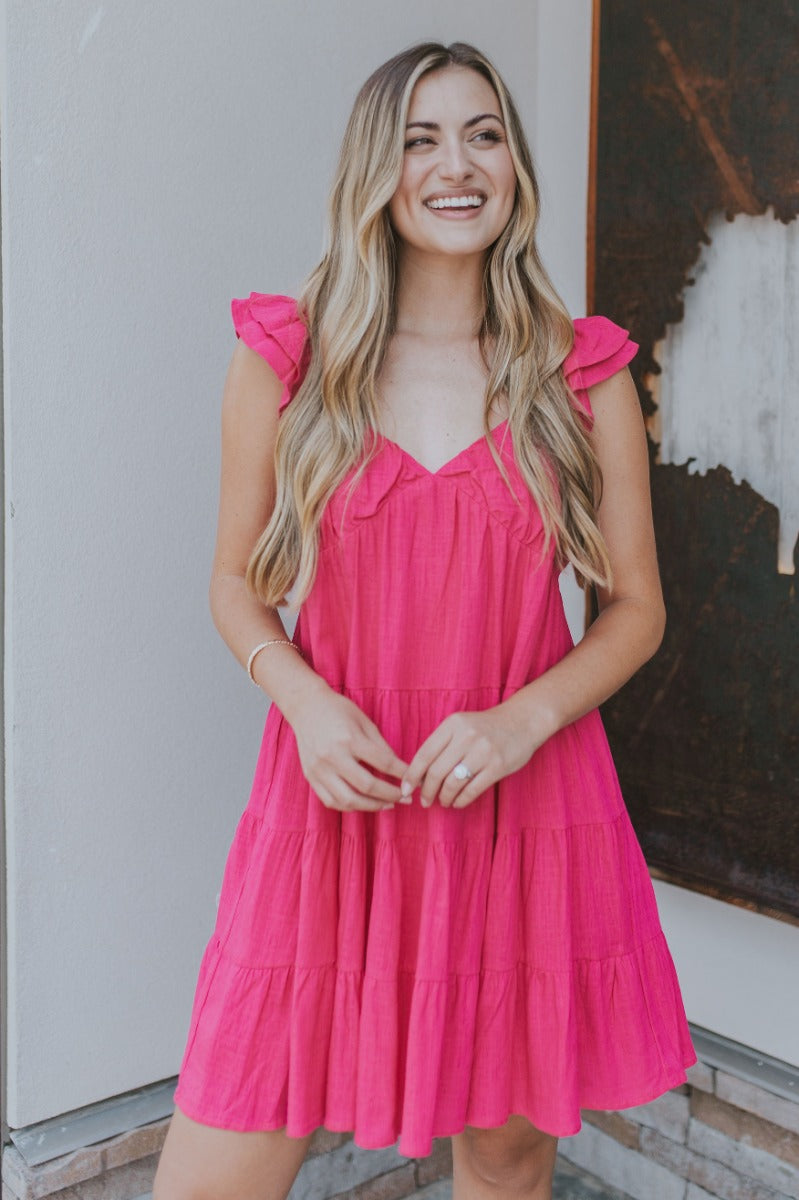 Front view of model wearing the Sweet Energy Mini Dress which features hot pink fabric, a mini hem length, a three-tiered body, hot pink lining, a v-neckline, and ruffle straps.