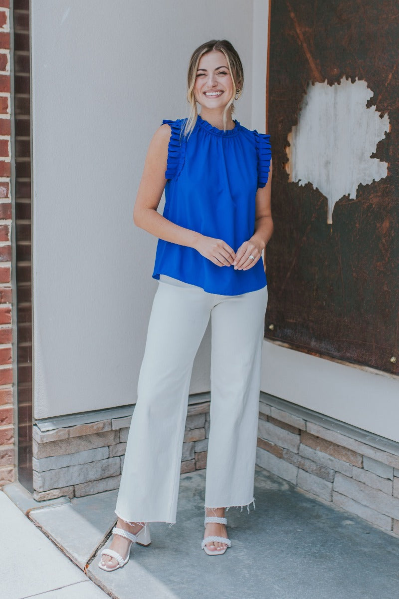 Full body front view of model wearing the Buena Vista Ruffle Tank that has royal blue fabric, a high neck with ruffle details, and a sleeveless design with pleated ruffle details