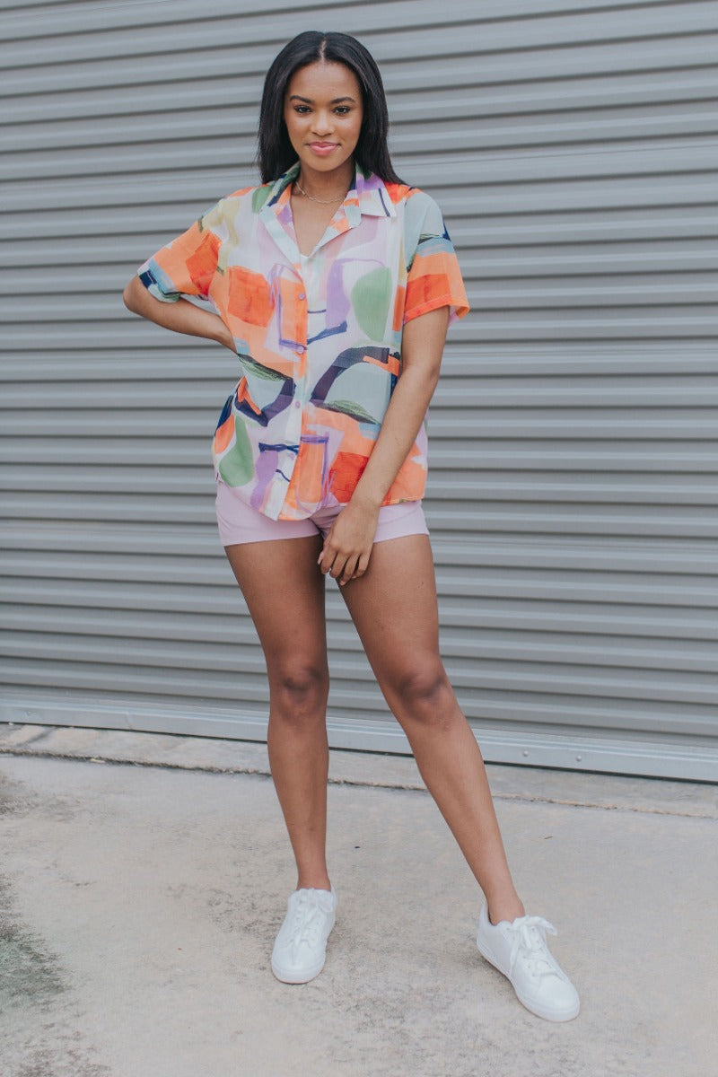 Full body view of model wearing the West Coast Top which features orange, purple, yellow, blue, green and white sheer fabric, geometric water color pattern, purple button down for closure, collared neckline and short sleeves.