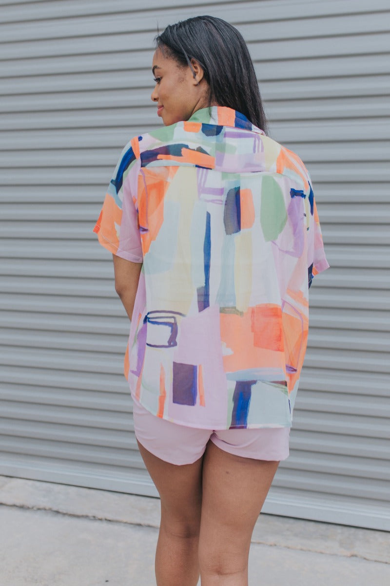 Back view of model wearing the West Coast Top which features orange, purple, yellow, blue, green and white sheer fabric, geometric water color pattern, purple button down for closure, collared neckline and short sleeves.