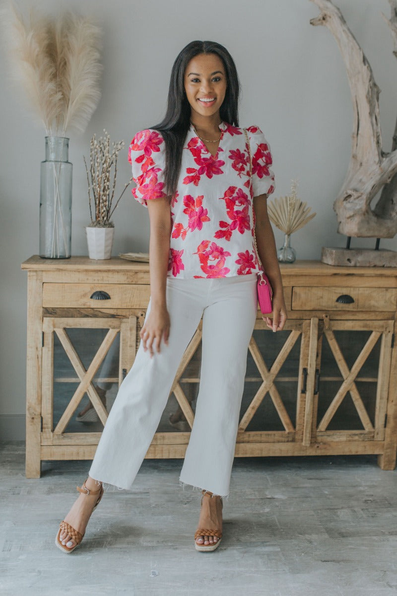 Full body view of model wearing the Nothing Compares Floral Top which features white brocade fabric with a fuchsia and red floral print, white lining, round neckline with a v-cutout and short puff sleeves.