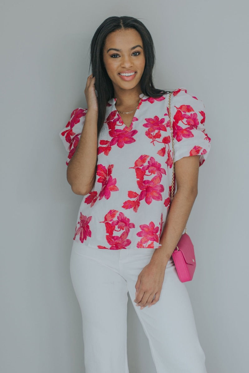 Front view of model wearing the Nothing Compares Floral Top which features white brocade fabric with a fuchsia and red floral print, white lining, round neckline with a v-cutout and short puff sleeves.