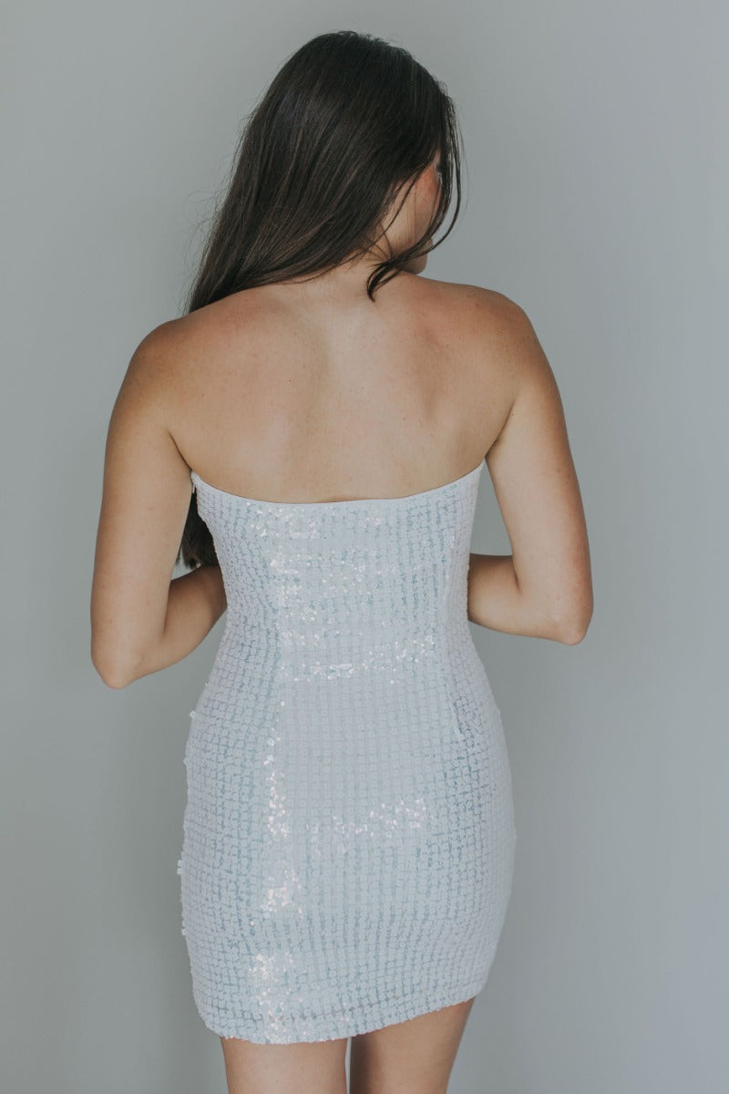 Back view of model wearing the Lost In A Dream Sequin Dress which features white square sequins, iridescent sequins, white lining, mini length, strapless and side zipper with hook closure.