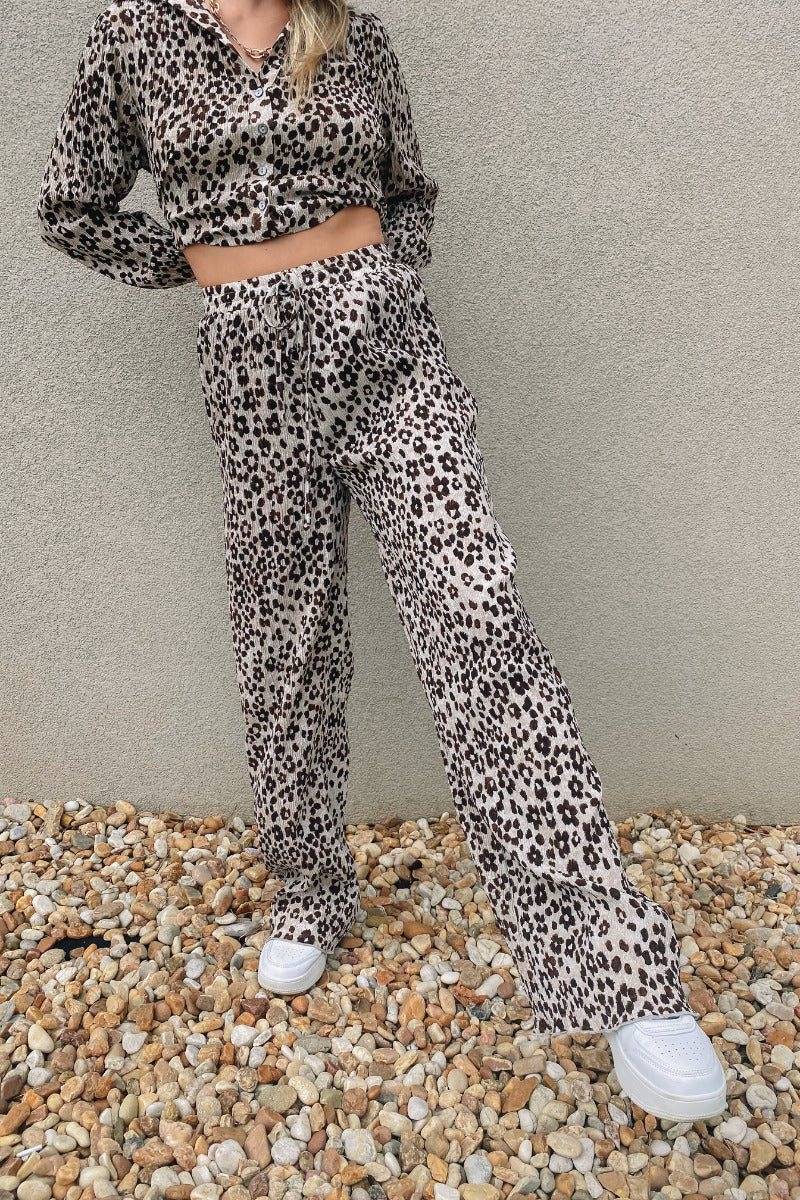 Frontal view of the Still Blooming Pants that features a taupe colored material, a high-rise fit, an elastic waist, adjustable ties at the waist, a lettuce edge hem, and a wide leg fit.