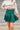 Frontal view of the In Harmony Skirt that features a hunter green material, a high-rise fit, a thick waist band, a pleated design throughout, and a back zipper closure