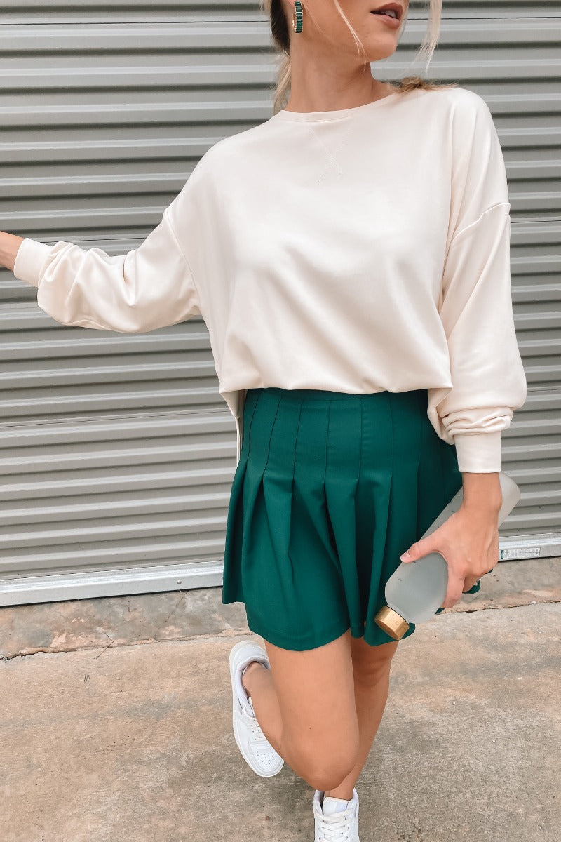Frontal view of the In Harmony Skirt that features a hunter green material, a high-rise fit, a thick waist band, a pleated design throughout, and a back zipper closure