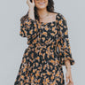 Front view of model wearing the Dare To Dream Floral Dress that has black lightweight fabric with rust florals, a bubble hem, mini length, upper boning, a square neck and 3/4 balloon sleeves.