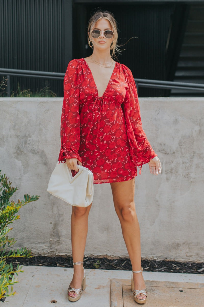 Full body front view of model wearing the Living The Good Life Dress in Red that has maroon sheer fabric with pink and red floral print, maroon lining, mini length, a plunge neck, an open back and long flare sleeves