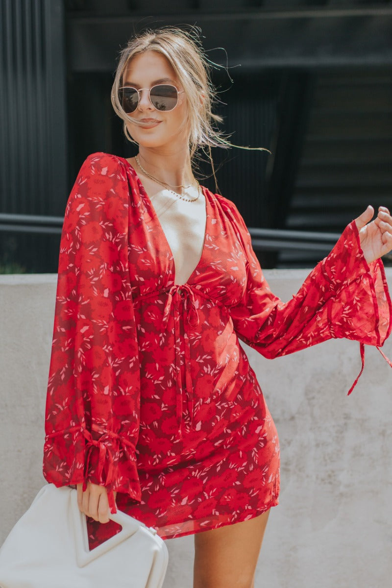 Front view of model wearing the Living The Good Life Dress in Red that has maroon sheer fabric with pink and red floral print, maroon lining, mini length, a plunge neck, an open back and long flare sleeves