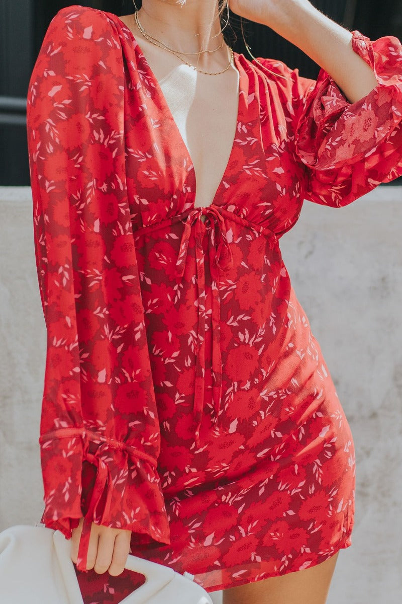 Close front view of model wearing the Living The Good Life Dress in Red that has maroon sheer fabric with pink and red floral print, maroon lining, mini length, a plunge neck, an open back and long flare sleeves