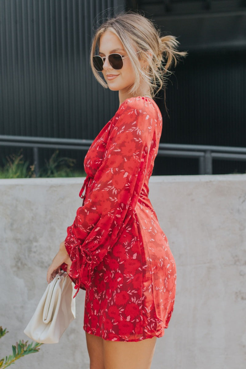 Side view of model wearing the Living The Good Life Dress in Red that has maroon sheer fabric with pink and red floral print, maroon lining, mini length, a plunge neck, an open back and long flare sleeves