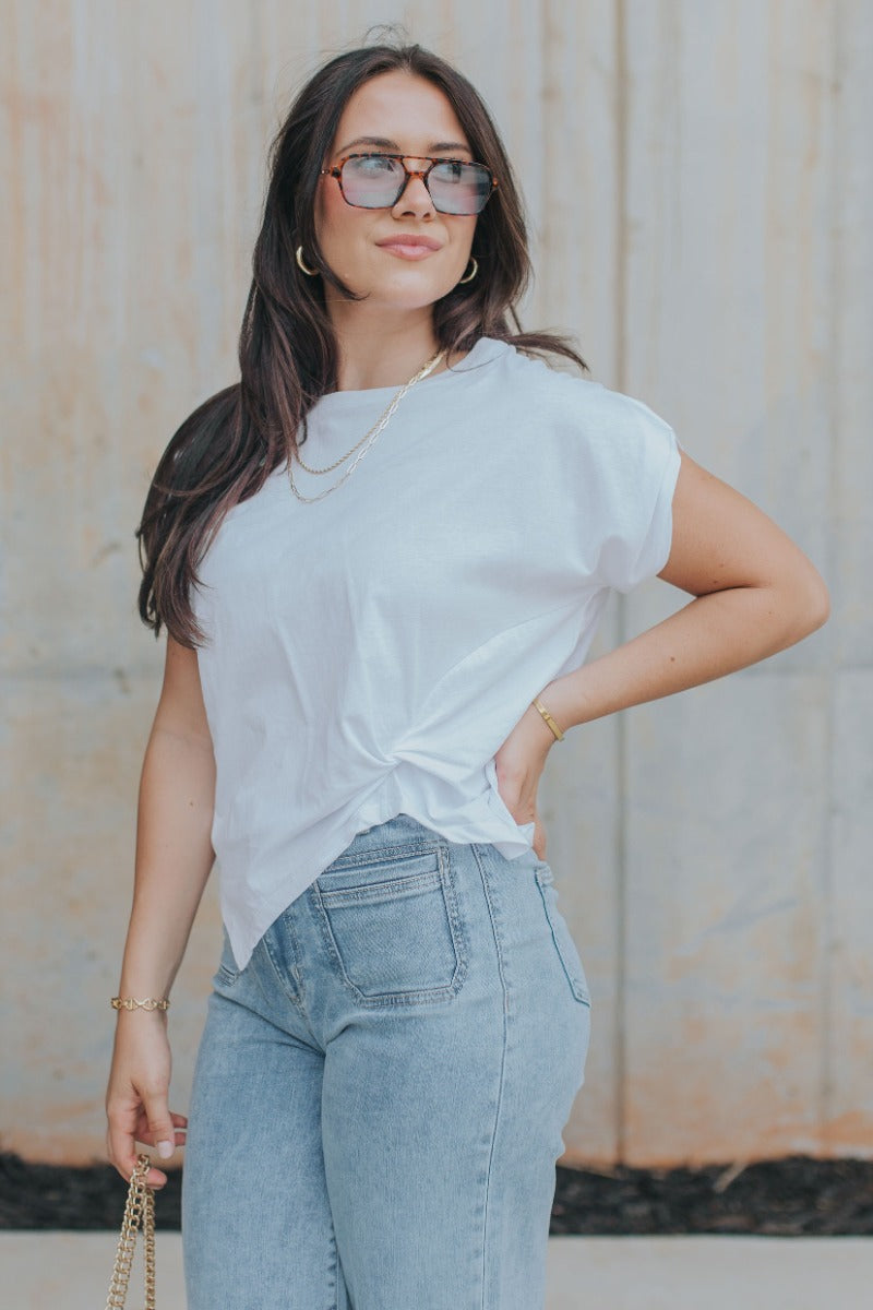 Frontal side view of model wearing the Tie The Knot Top which features white cotton fabric, side twist knot detail, round neckline and short sleeves.