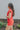 Side view of model wearing the Game On Romper which features washed red denim fabric, distressed details, two front pockets, one chest pocket, folded hem shorts, an elastic waistband in the back, a hidden zipper and button up closure, a collared neckline,