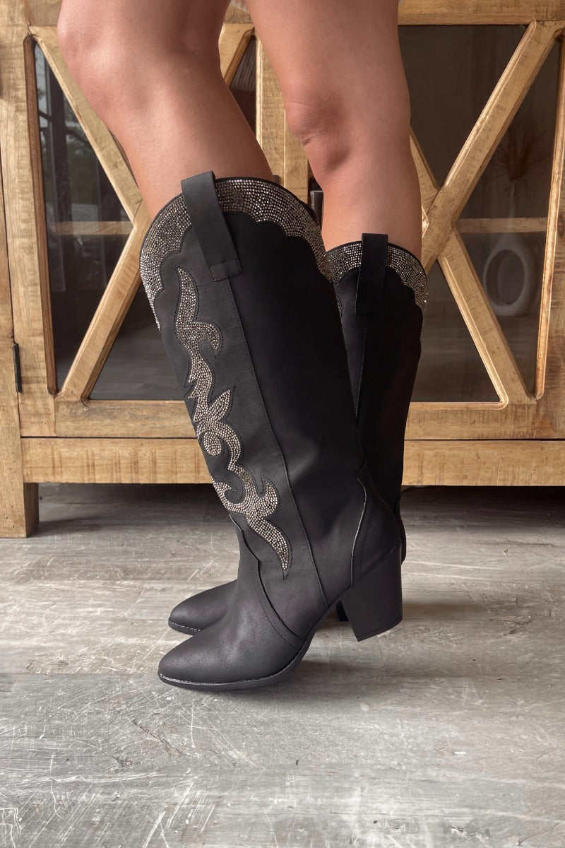Side view of model wearing the Zane Rhinestone Cowgirl Boot in Black which features a western style pattern with rhinestone details, tall boot, pull tabs, monochromatic heel and chic pointed toe.