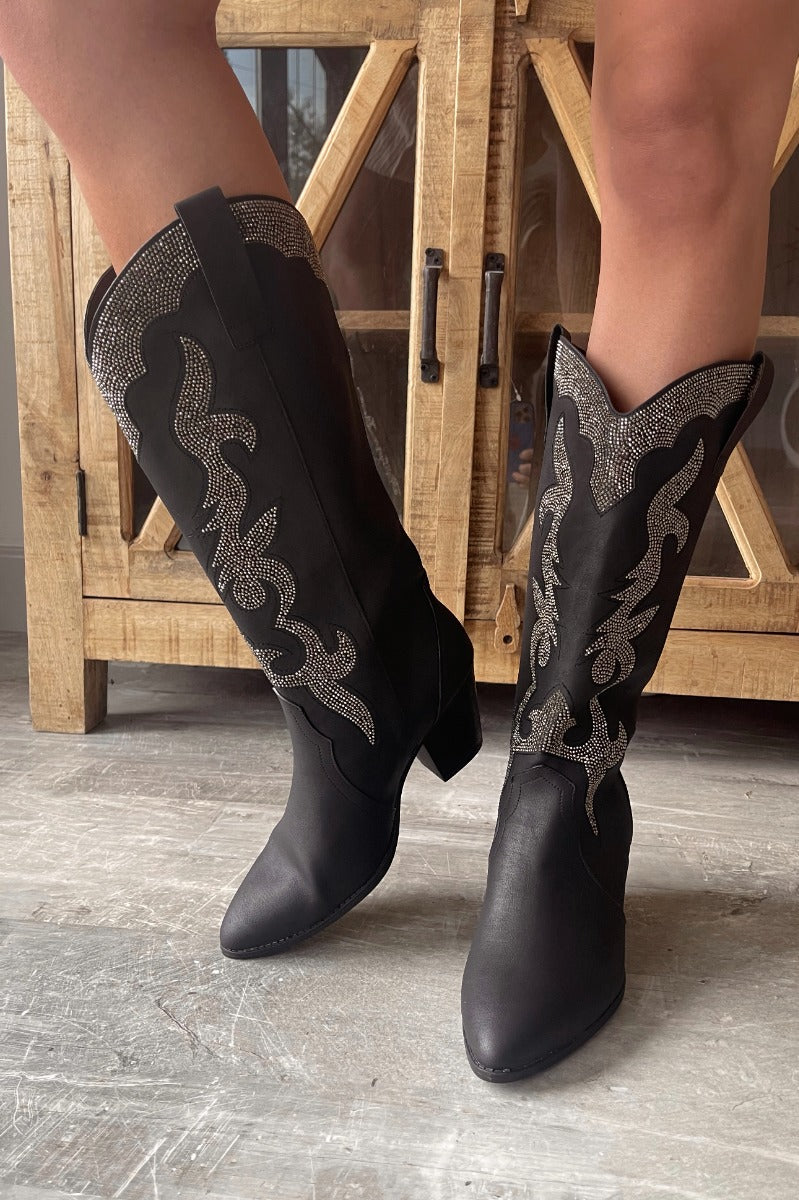 Front view of model wearing the Zane Rhinestone Cowgirl Boot in Black which features a western style pattern with rhinestone details, tall boot, pull tabs, monochromatic heel and chic pointed toe.