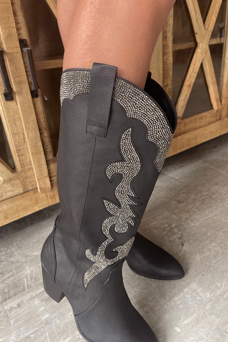 Close up view of model wearing the Zane Rhinestone Cowgirl Boot in Black which features a western style pattern with rhinestone details, tall boot, pull tabs, monochromatic heel and chic pointed toe.