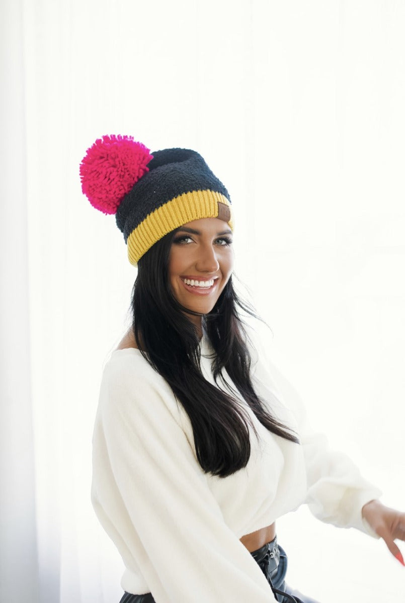 Frontal view of model wearing the Stay Cozy Beanie in Black, that features black sherpa fabric, yellow trim, and a pink pom pom.