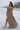 Image shows brunette female model on beach. Model is wearing a brown maxi dress.