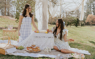 Image shows two women at a picnic. They are kneeling at a table with food. Both women are wearing white printed dresses.