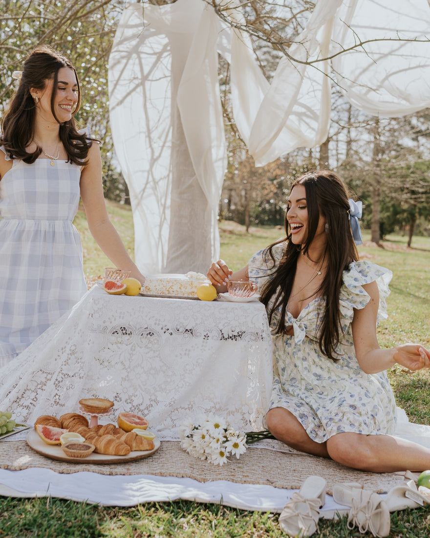 Image shows two women at a picnic. They are kneeling at a table with food. Both women are wearing white printed dresses.