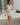 Image shows full body front view of female model wearing a white mini dress and holding a beige purse.