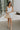 Image shows full body front view of female model wearing a white mini dress and holding a beige purse.