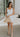 Image shows full body front view of female model wearing a white mini dress and holding a beige purse.
