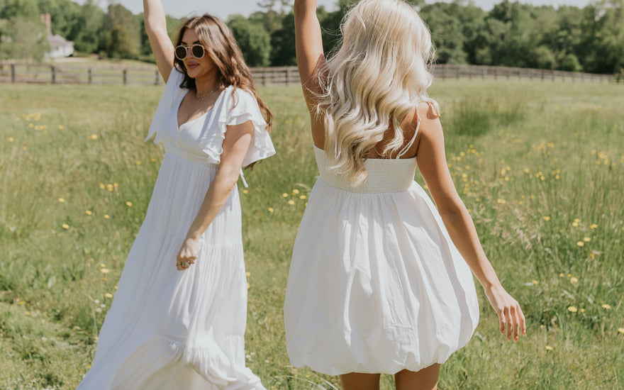 Image shows 2 young women outside in field. left woman is wearing a white maxi dress and left woman has her back turned and is wearing a white mini dress.